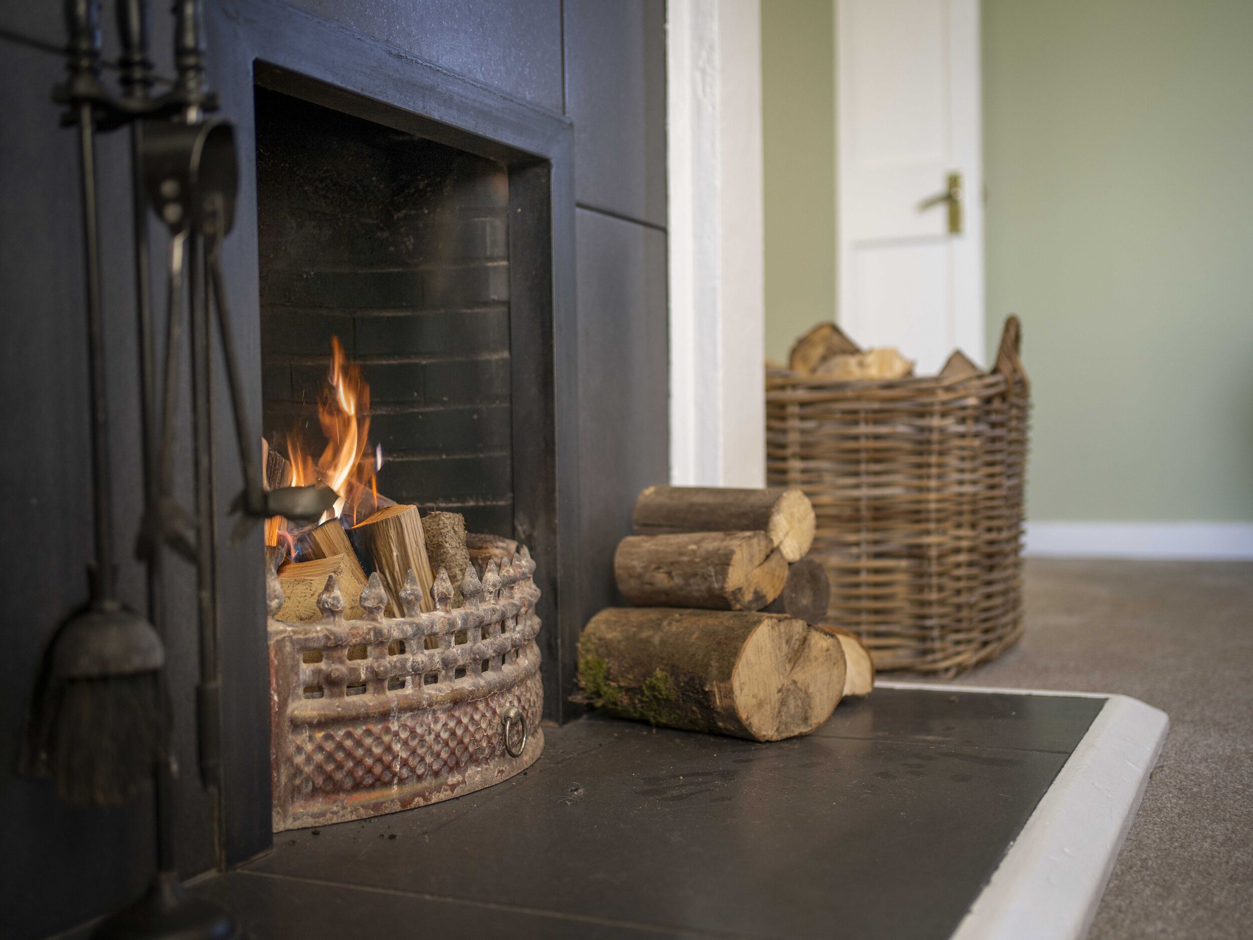 A fireplace with a fire burning logs, a metal grate, and a decorative metal piece. There are wicker baskets with logs and a door in the background.