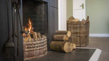 A fireplace with a fire burning logs, a metal grate, and a decorative metal piece. There are wicker baskets with logs and a door in the background.