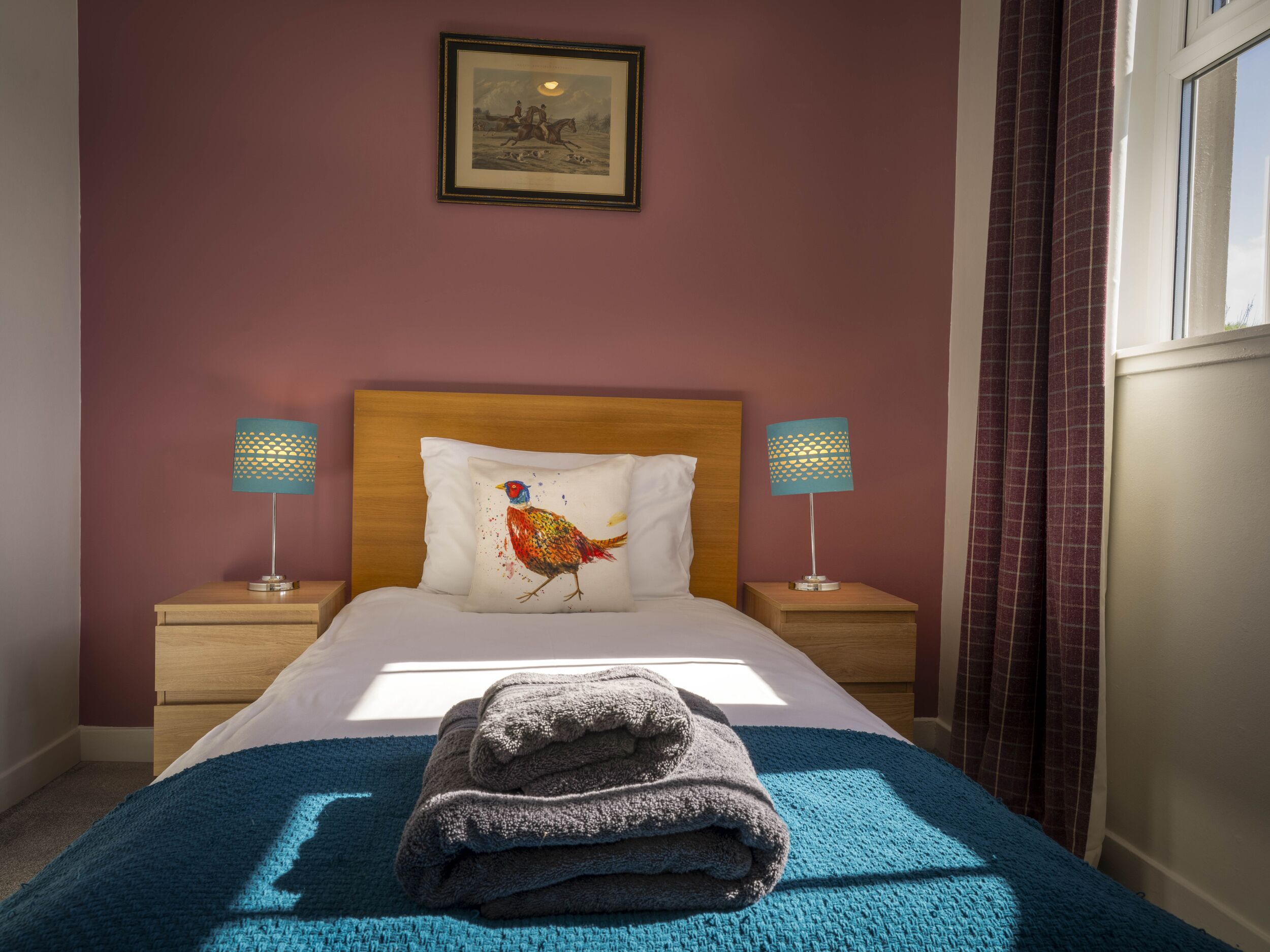 Un lit bien fait, recouvert d’une couverture bleue, avec un oreiller orné d’un motif coloré représentant un oiseau, et deux lampes de chevet aux abat-jour à motifs.