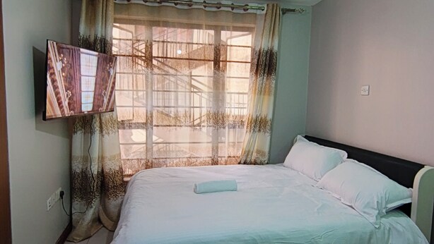 A bedroom with a bed, a TV mounted on the wall, and a window with curtains. - Beautiful Studio apartment in Nairobi near JKIA/SGR perfect for your stay (Nairobi)