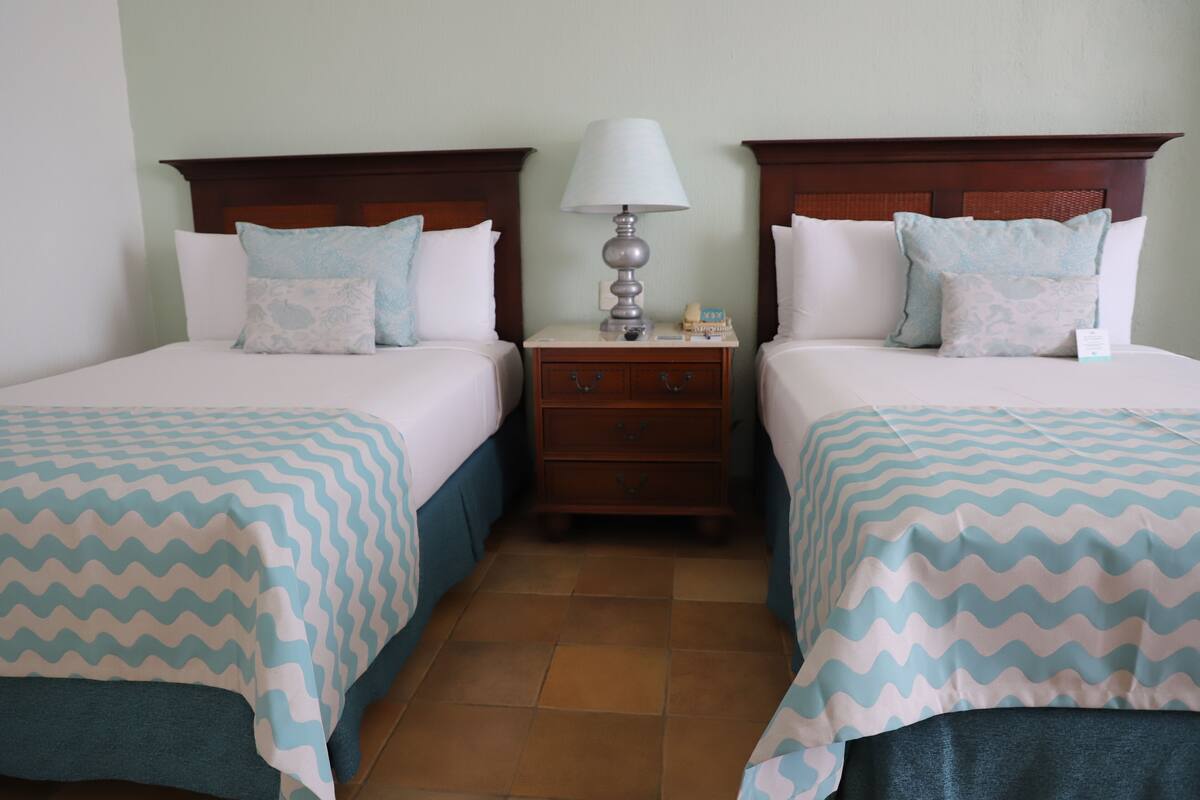 A hotel room with two beds, each with a patterned bedspread, a nightstand with a lamp, and a small drawer.