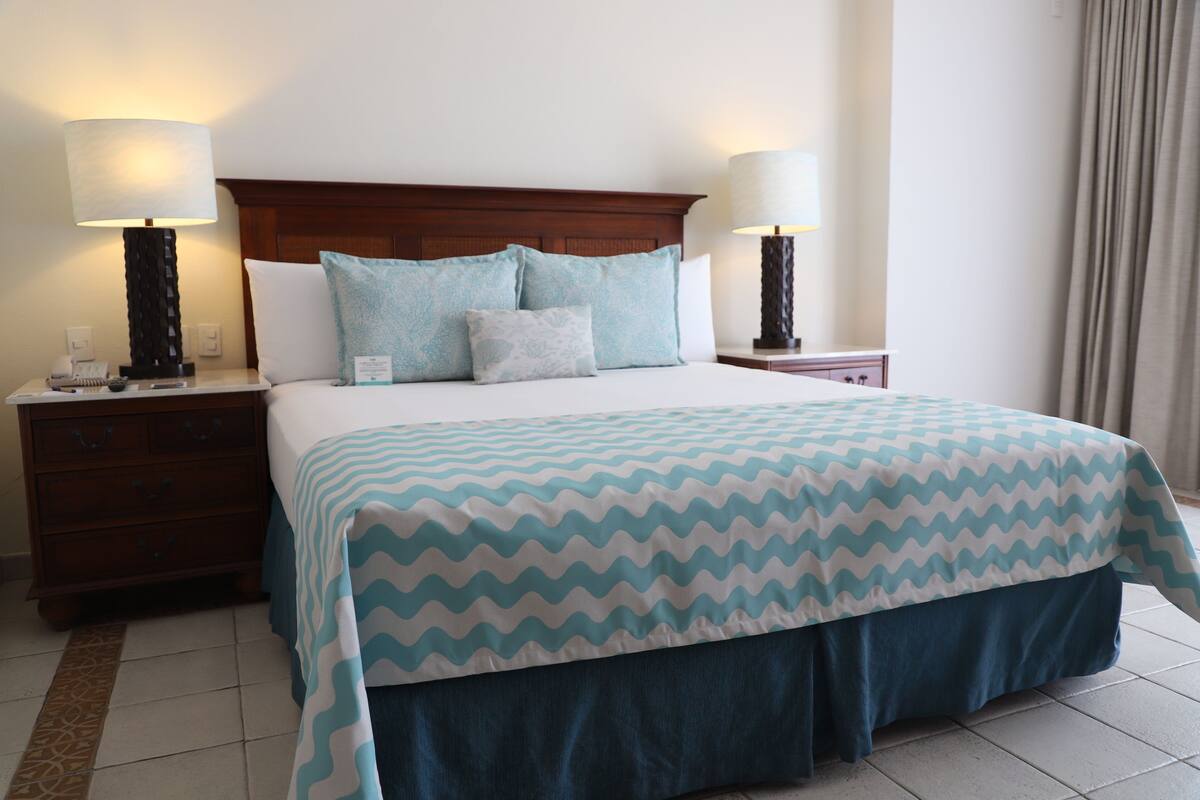A hotel room with a bed, two bedside tables, lamps, a wooden headboard, and a patterned bedspread.