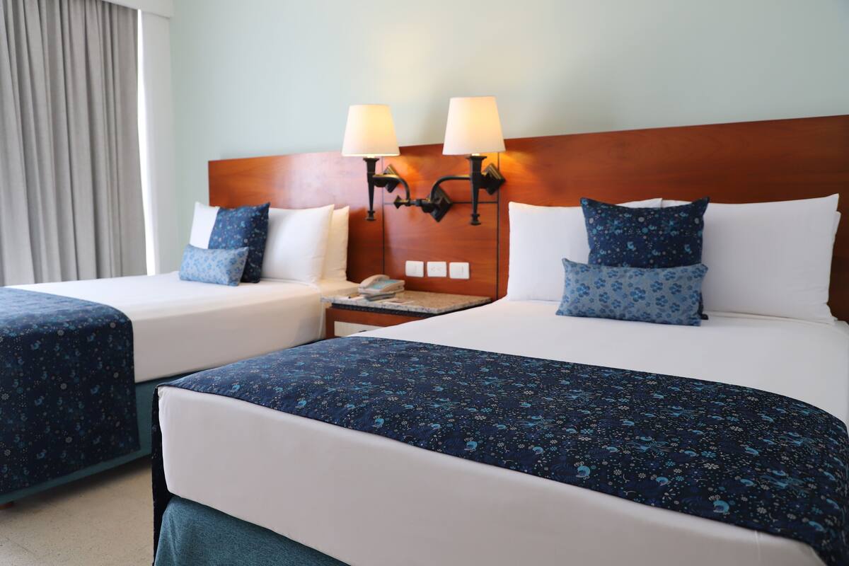A hotel room with two beds, each with a blue floral bedspread, white pillows, and a wooden headboard. There are two wall-mounted lamps on the headboard and a nightstand with a phone and a lamp.