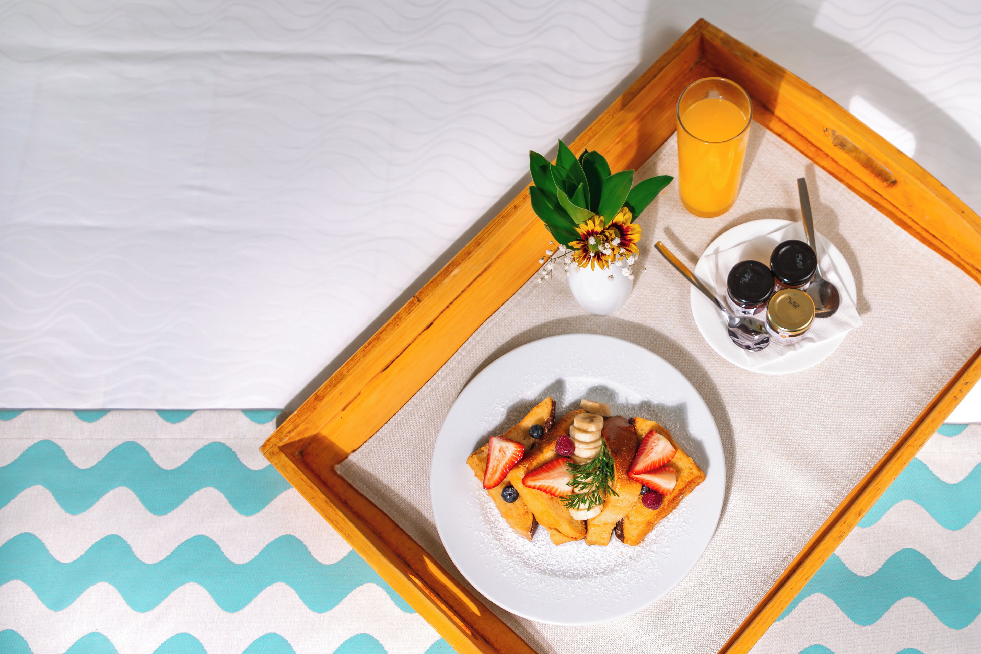 Een ontbijtkarretje met pannenkoeken, aardbeien, bosbessen en een glas sinaasappelsap op een bed met een blauw-witte sprei.