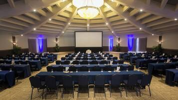 Une salle de conférence avec des rangées de tables et de chaises, un grand écran et un lustre.