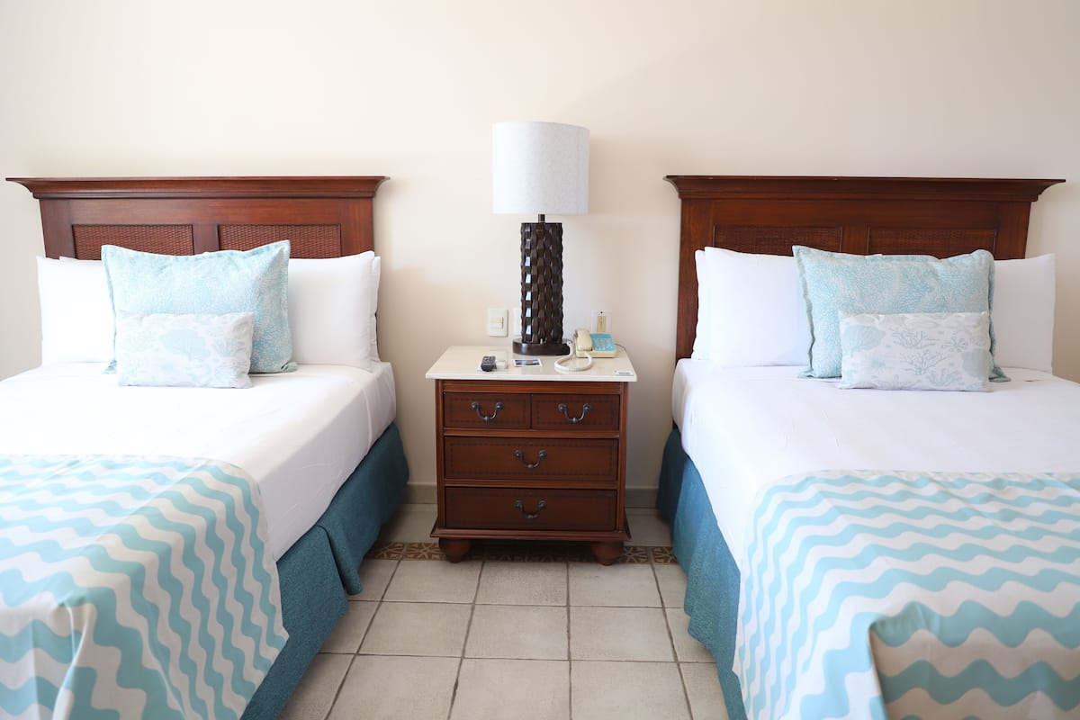 A hotel room with two beds, each with a blue and white zigzag patterned bedspread, a wooden nightstand with a lamp and phone, and a tiled floor.