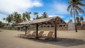 A beach with a wooden structure providing shade, several lounge chairs, and palm trees in the background. - Pierre Mundo Imperial All Inclusive  (Acapulco)