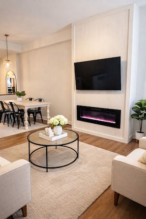 A modern living room with a flat-screen TV mounted on a wall, a fireplace below it, a round coffee table with a vase of flowers, and a dining area in the background.