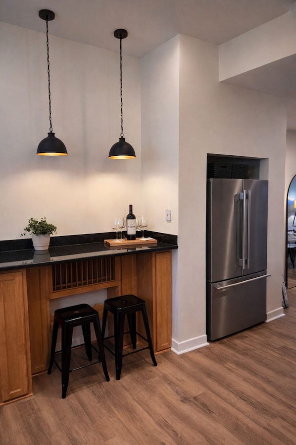 A kitchen with a stainless steel refrigerator, wooden bar counter, two black pendant lights, and a potted plant.