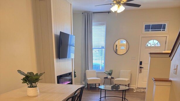 Uma sala de jantar com uma mesa branca e cadeiras pretas, uma pequena mesa redonda com uma planta e um ventilador de teto.