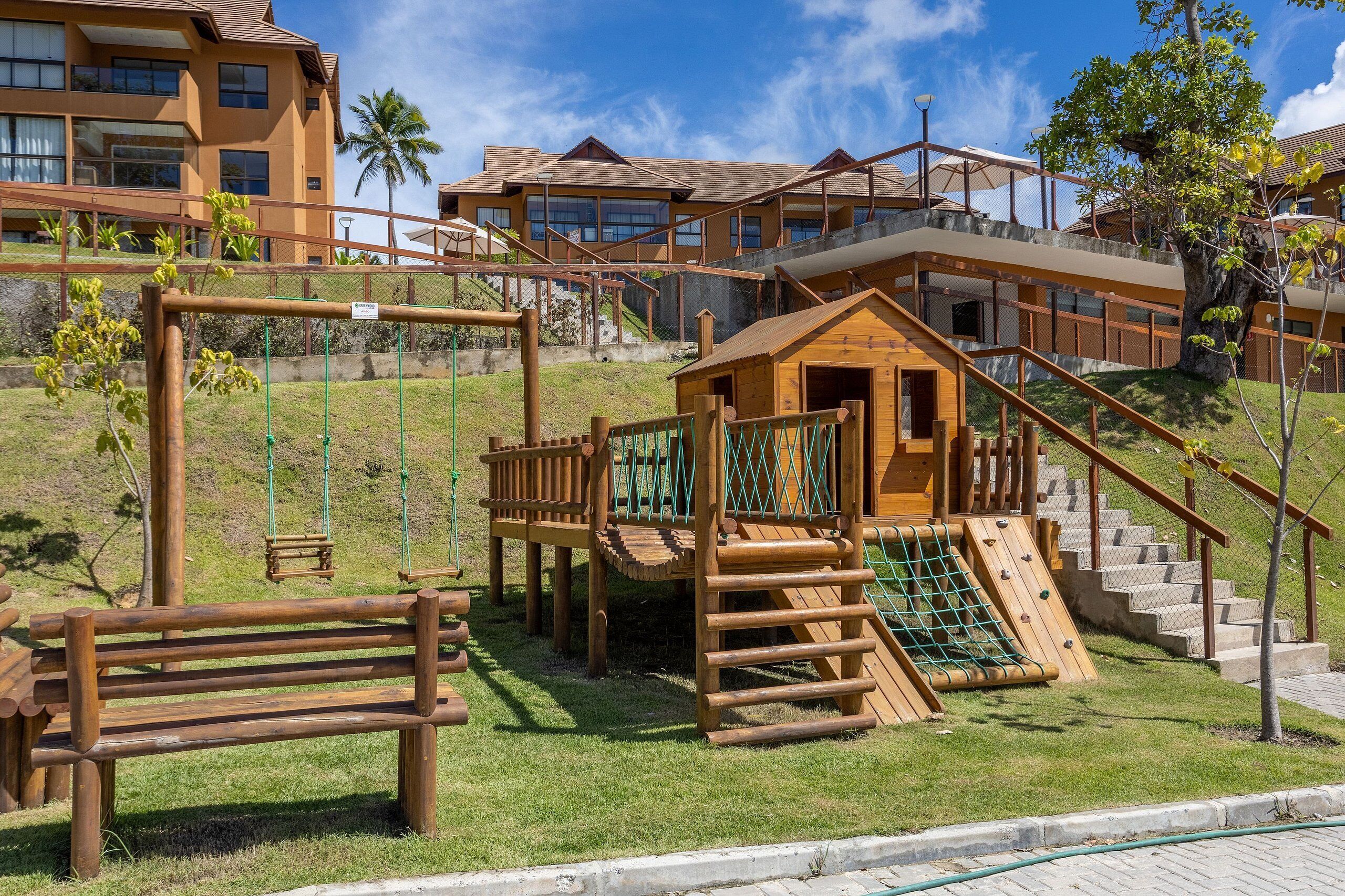 A playground with wooden structures, including a swing set and a climbing area, surrounded by a grassy area and a building in the background.