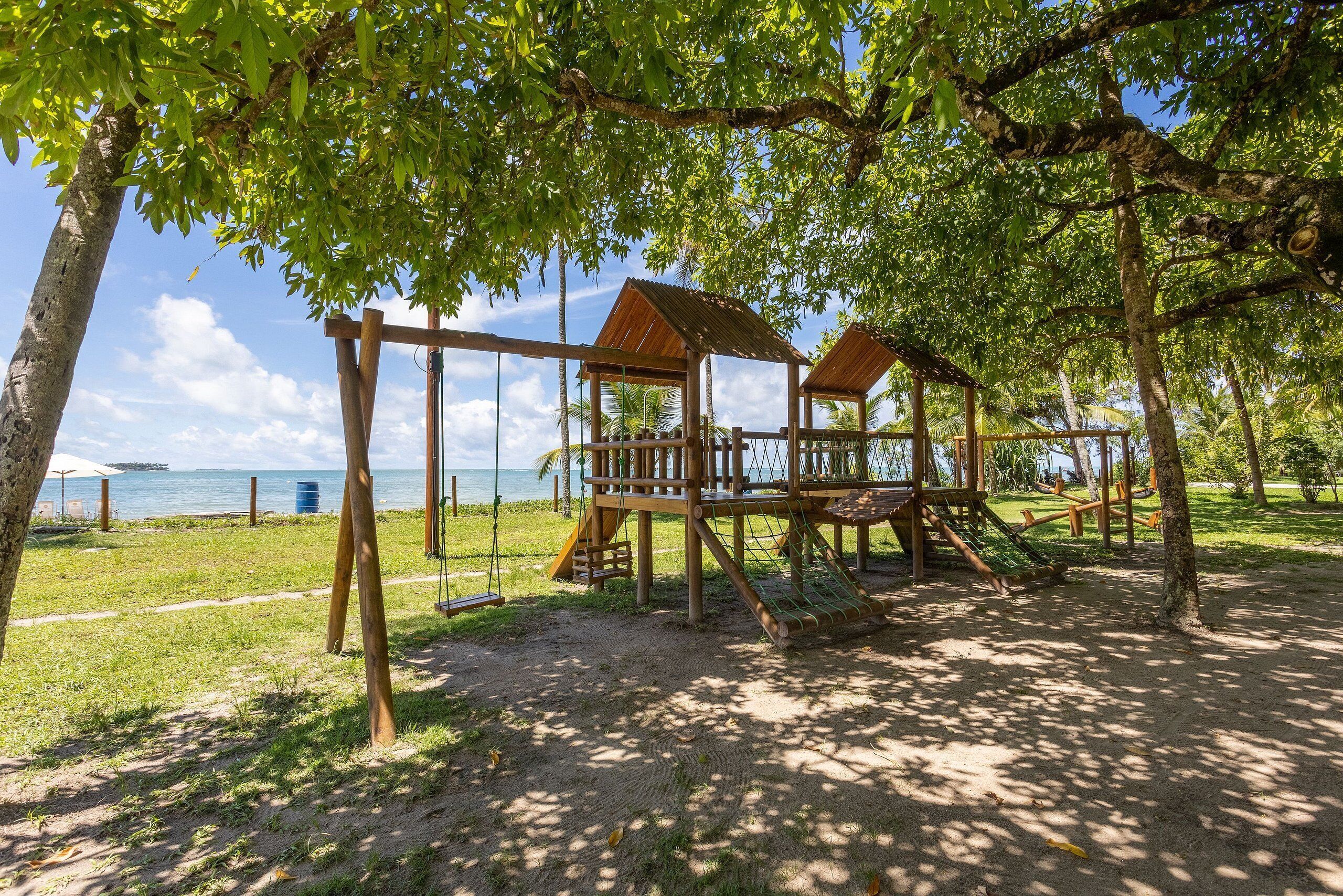Un parque infantil de madera con columpios y toboganes, rodeado de árboles y vistas al mar.