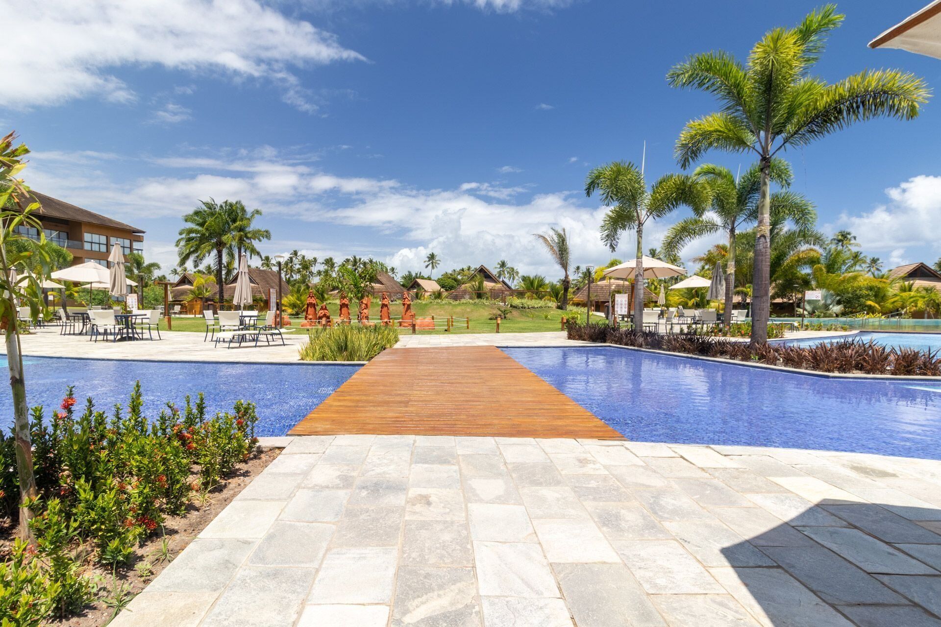 A resort pool area with a wooden walkway, palm trees, and lounge chairs.