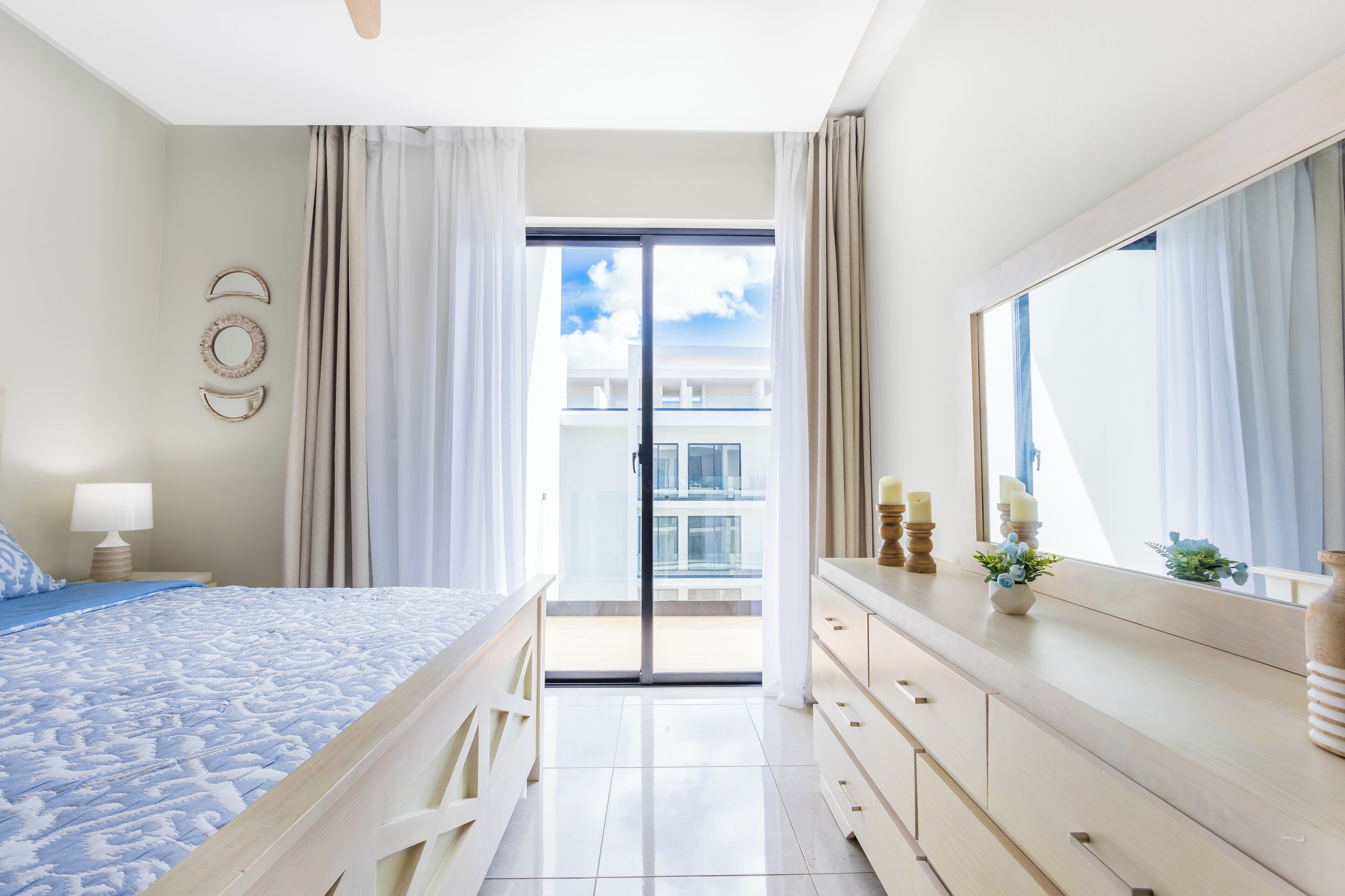 Une chambre d’hôtel comprenant un lit, une commode, un miroir, une table de chevet et donnant sur un balcon et un immeuble.