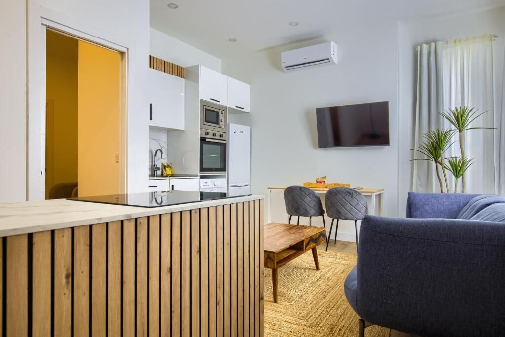 A modern kitchen with white cabinets, a built-in oven, and a microwave. There is a dining area with a wooden table and two chairs. A flat-screen TV is mounted on the wall, and a plant is visible in the corner.