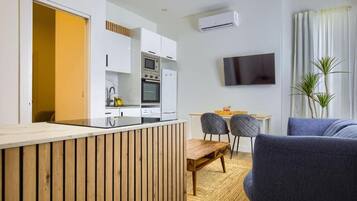 Una cocina moderna con armarios blancos, horno empotrado y microondas. Hay una zona de comedor con una mesa de madera y dos sillas. Un televisor de pantalla plana está montado en la pared, y se ve una planta en la esquina.