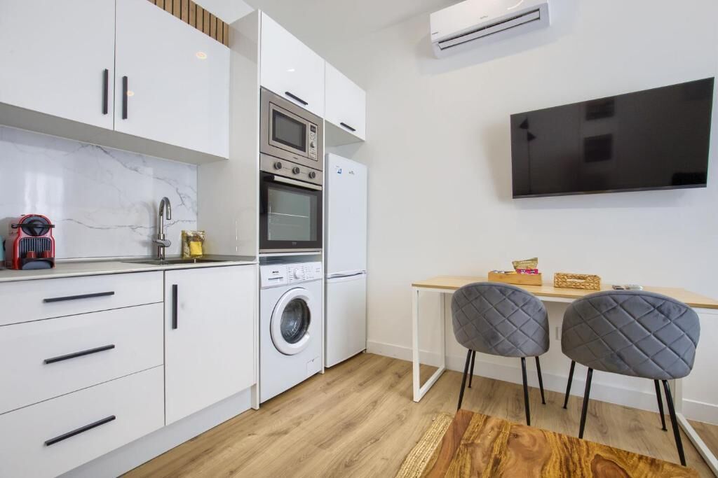 Une cuisine moderne avec des meubles blancs, un four encastré, un micro-ondes et un lave-linge. On y trouve une table à manger en bois avec deux chaises, ainsi qu’un téléviseur à écran plat fixé au mur.