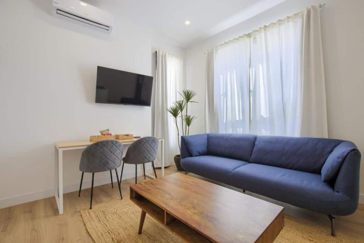 A modern living room with a blue sofa, a wooden coffee table, two chairs, a TV mounted on the wall, and a large window with sheer curtains.