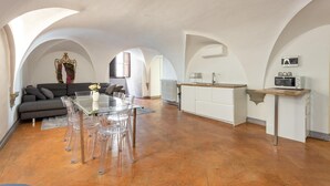 Un vaste espace de vie comprenant un coin repas avec une table en verre, un canapé gris et une cuisine dotée d’un plan de travail en bois et de meubles blancs.