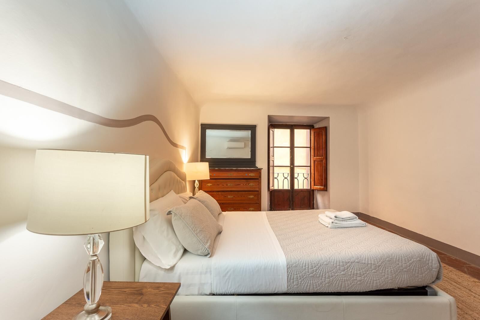 A hotel room with a bed, a nightstand, a lamp, a dresser, and a door with a window.