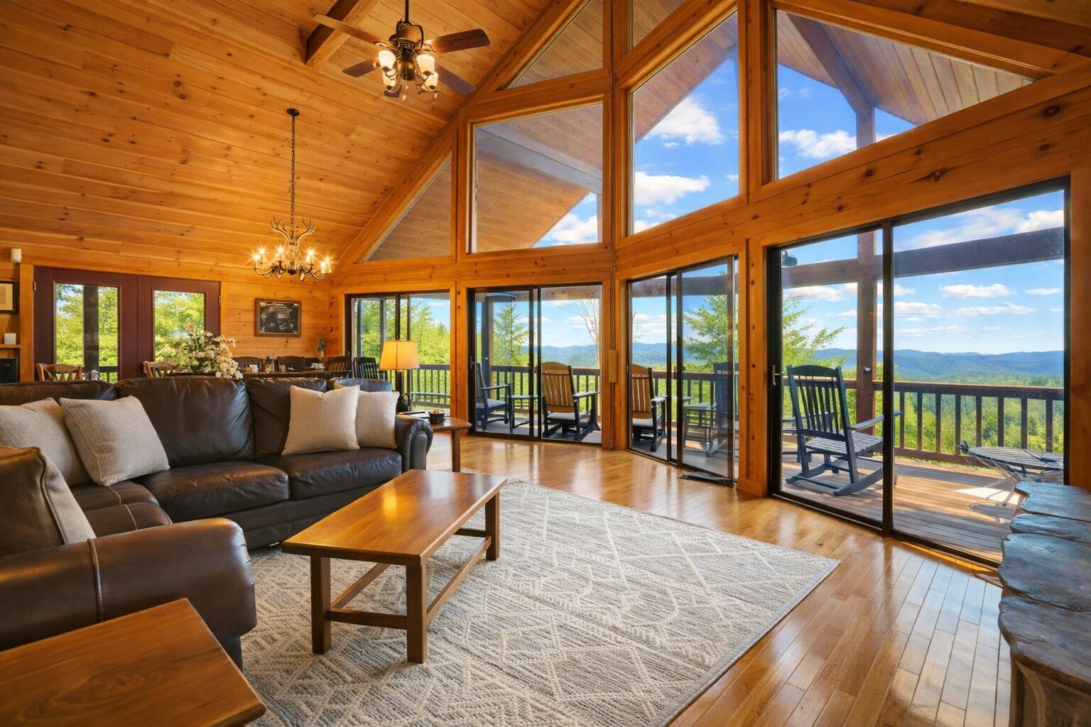 A spacious living room with a leather sofa, wooden coffee table, and a chandelier. Large windows offer a view of the outdoors.