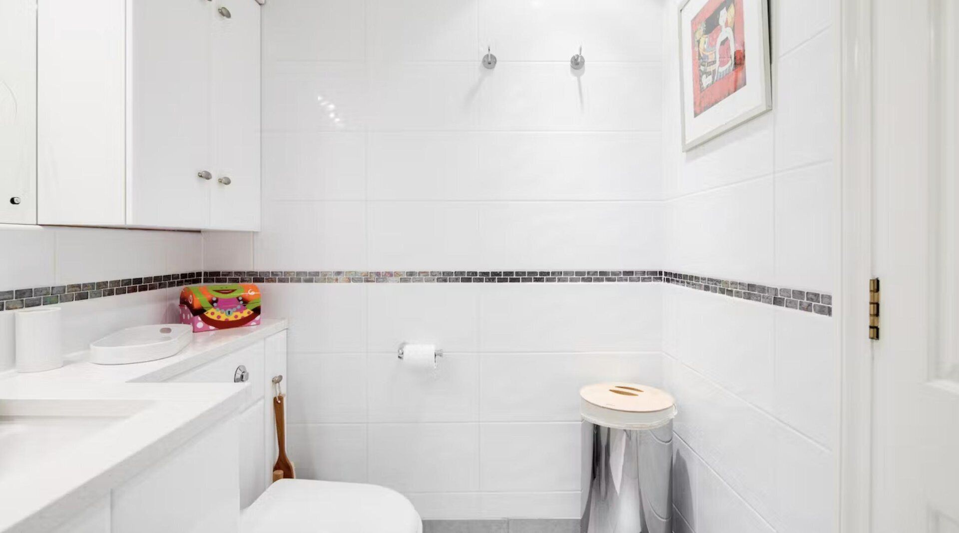 A bathroom with white tiles, a toilet, a sink, a trash bin, and a shower.