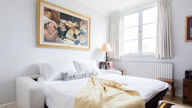 Una camera d'albergo con un letto, un comodino, una lampada, una finestra con tende e un quadro appeso al muro.