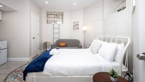 A bedroom with a bed, a desk, a chair, a small table, a plant, a ladder, and a window with blinds. - Petit Studio in Back Bay (Boston)