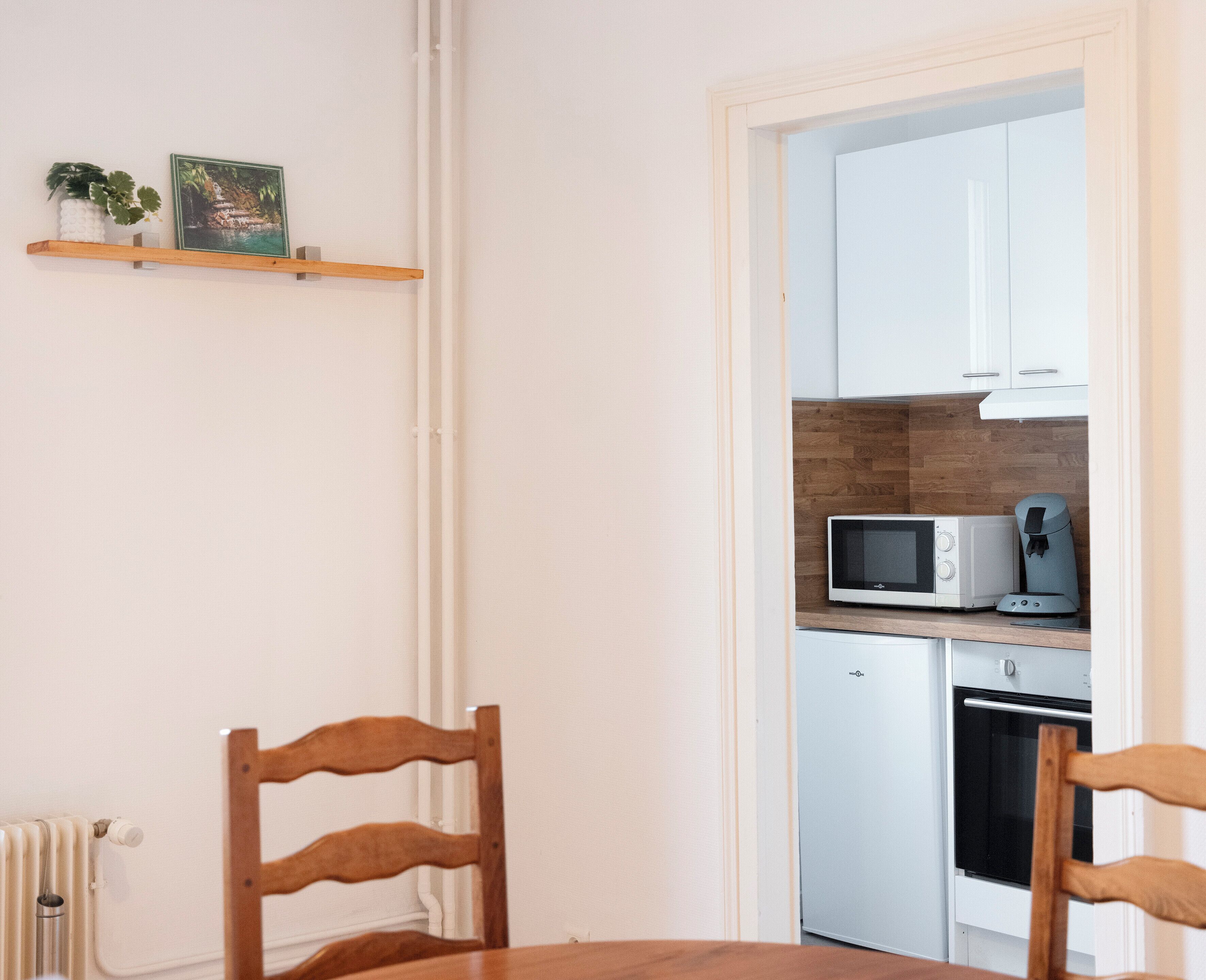 A compact kitchen with white cabinets, a microwave, and a coffee maker. There is a wooden chair at the table and a radiator on the wall.