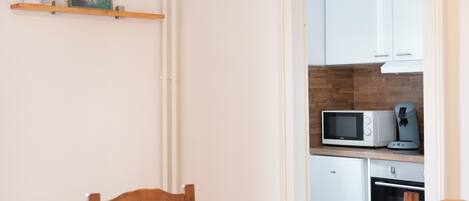 A compact kitchen with white cabinets, a microwave, and a coffee maker. There is a wooden chair at the table and a radiator on the wall.