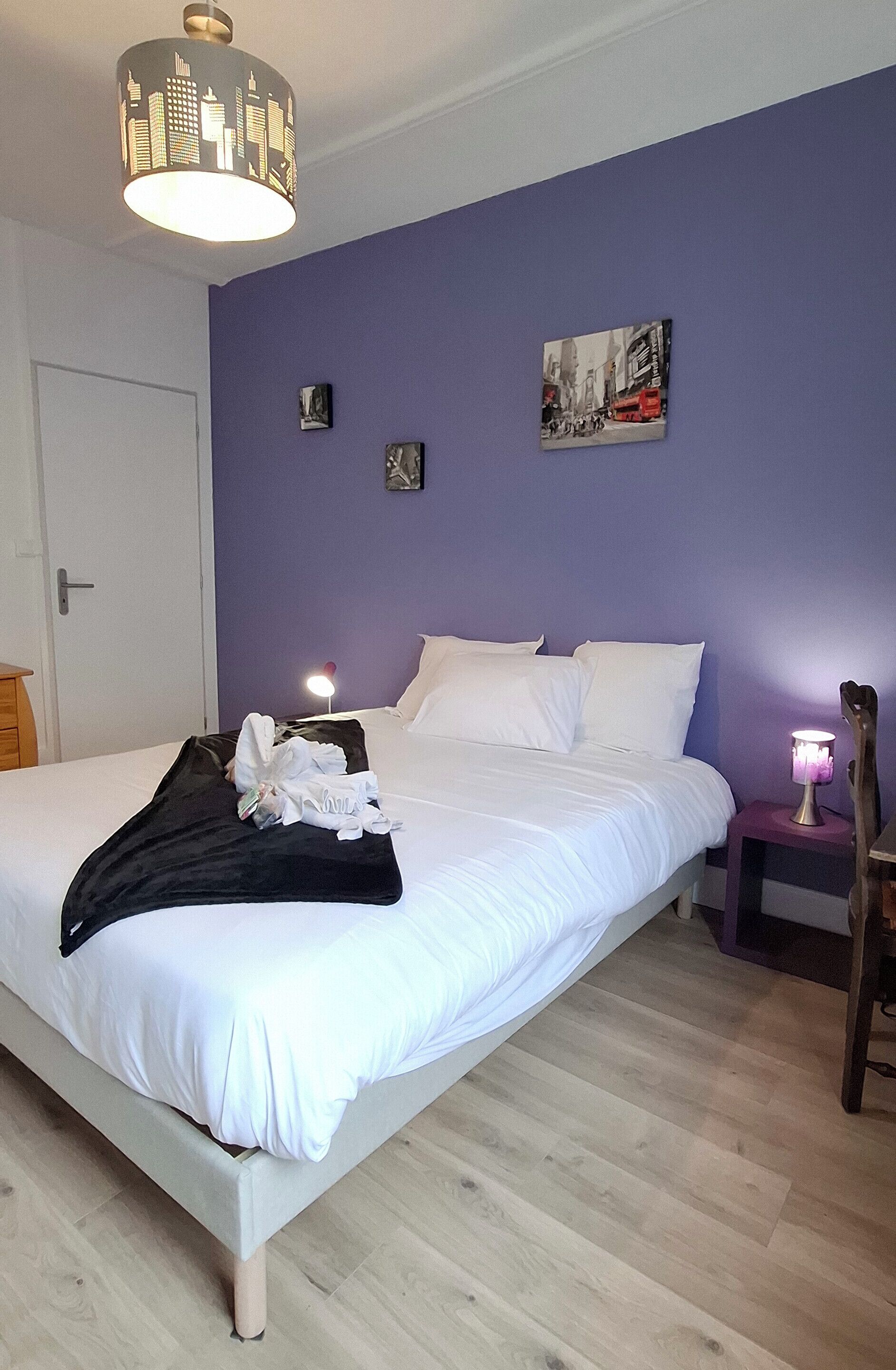 Letto rifatto con lenzuola bianche, una coperta nera e un piccolo mucchio di asciugamani piegati sul lato. All'interno, un'opera d'arte che raffigura una veduta urbana, un comodino con una lampada e una porta a sinistra.