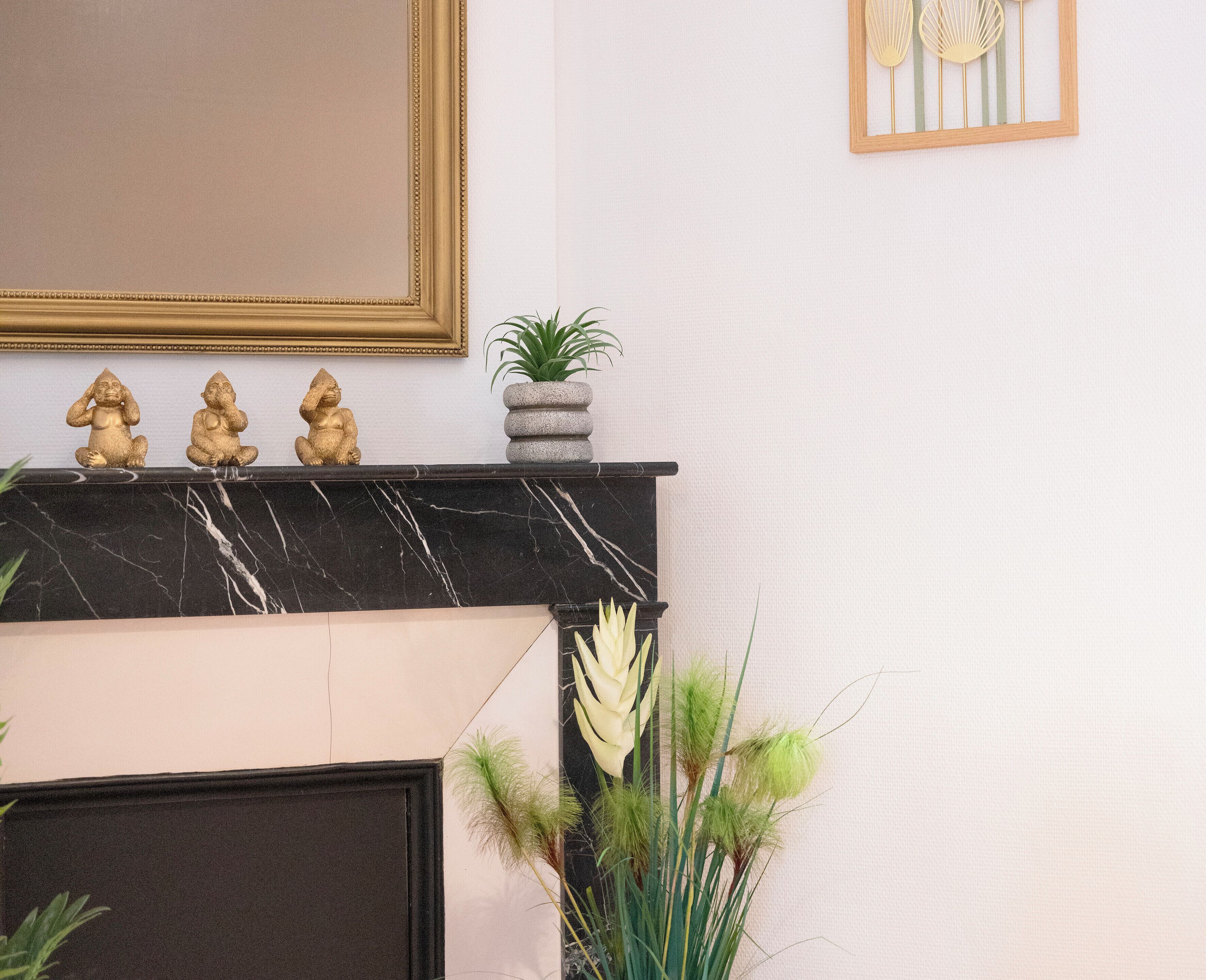 Een open haard met een marmeren bovenplaat, drie gouden beelden, een kleine potplant en decoratieve wandkunst.