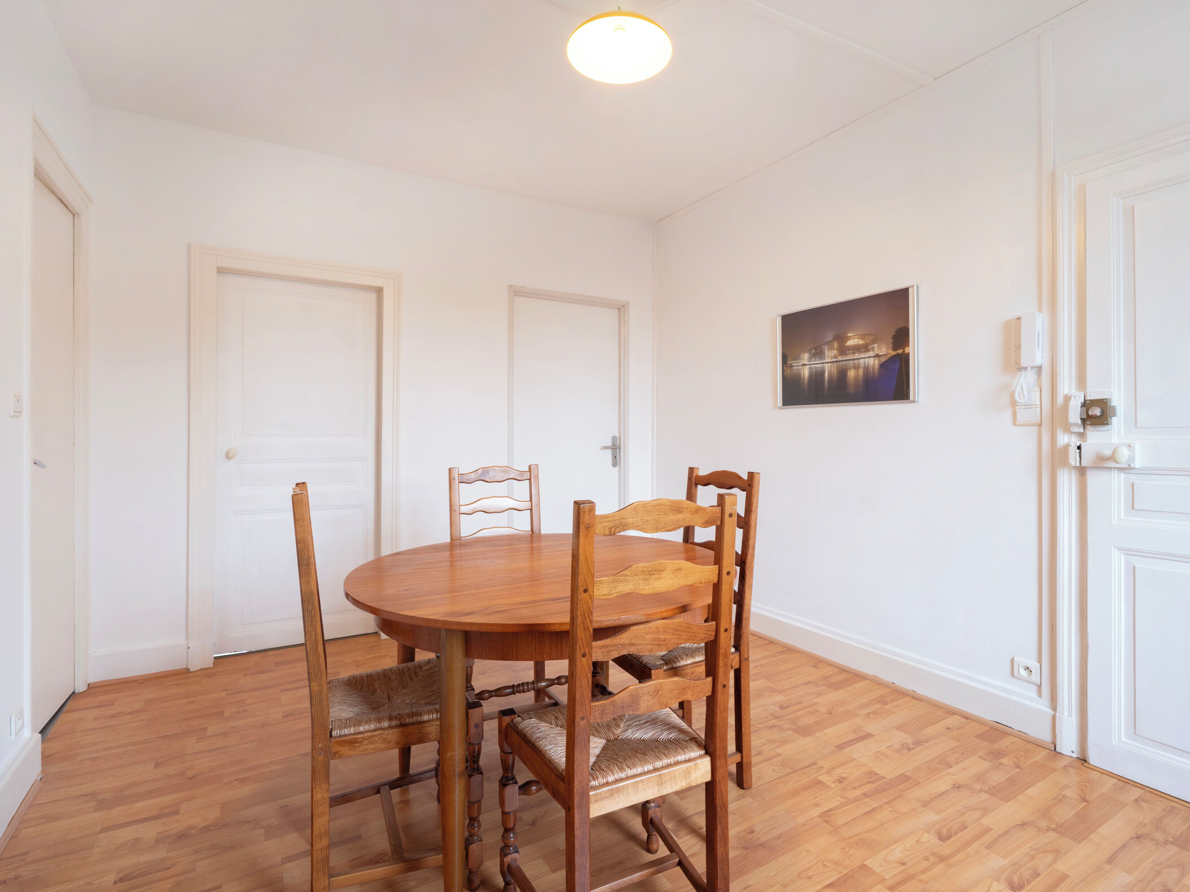 A dining area with a round wooden table and four chairs, white walls, and a ceiling light.