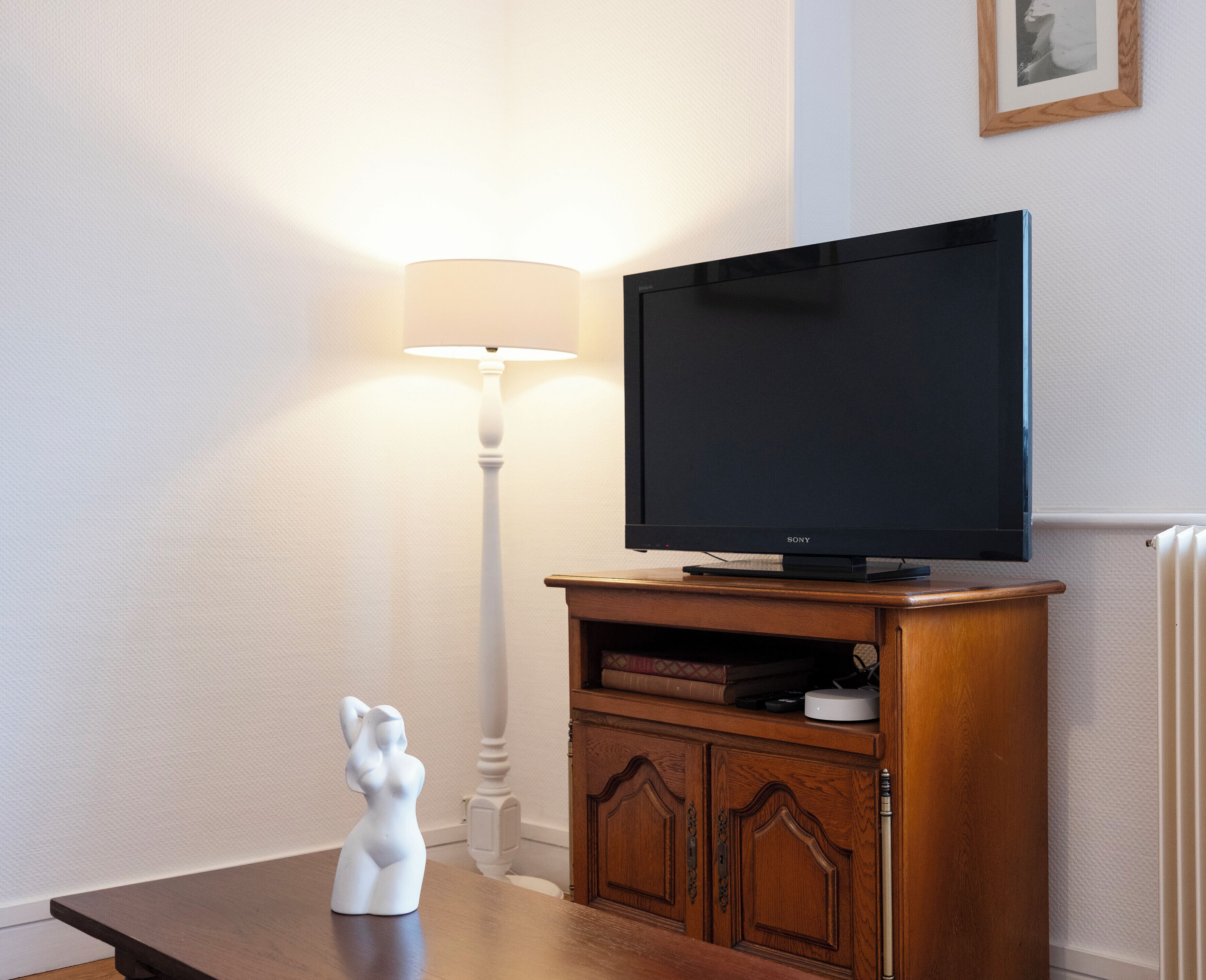 Ein Wohnzimmer mit Flachbildfernseher auf einem Holzständer, einer weißen Stehlampe und einer kleinen Statue auf einem Holztisch.