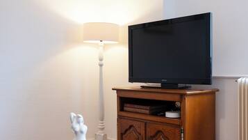 Ein Wohnzimmer mit Flachbildfernseher auf einem Holzständer, einer weißen Stehlampe und einer kleinen Statue auf einem Holztisch.