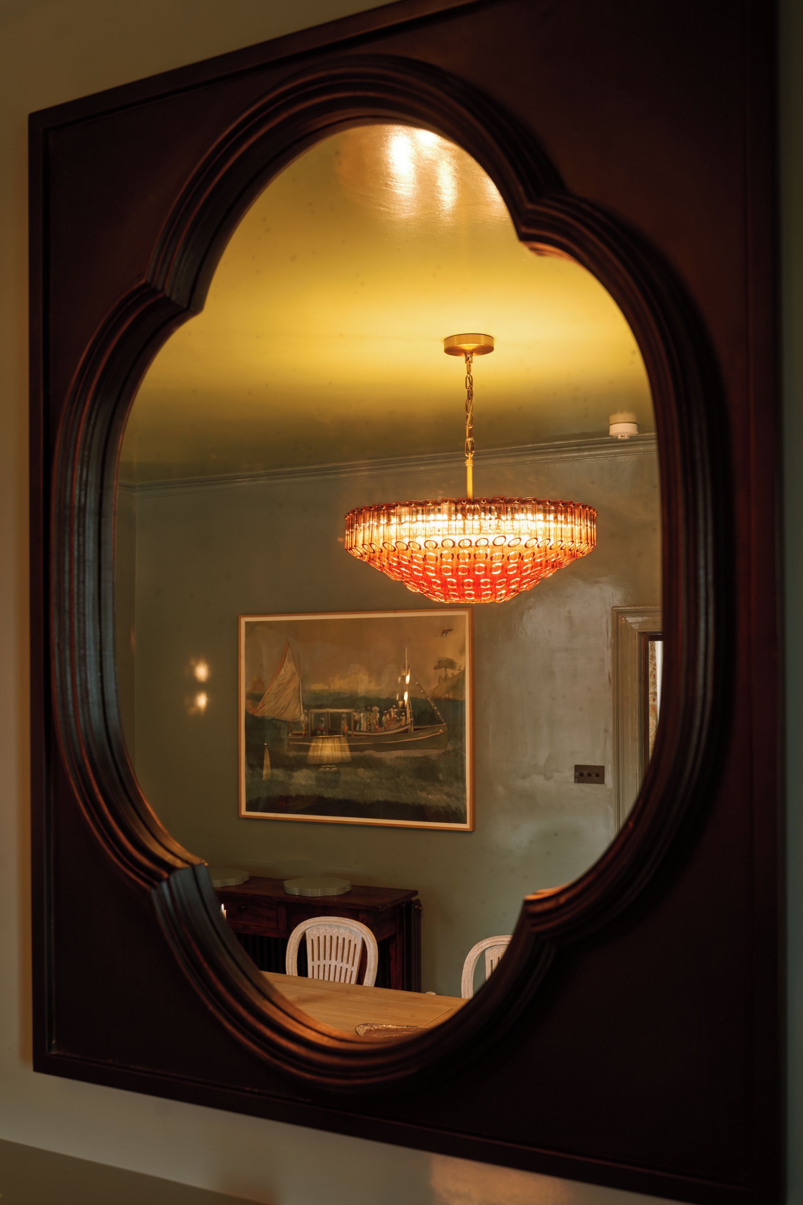 A mirror with a decorative frame reflecting a chandelier and a dining area.