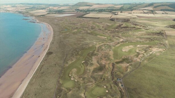 An aerial view of a coastal golf course with a beach, green fairways, and sand traps.