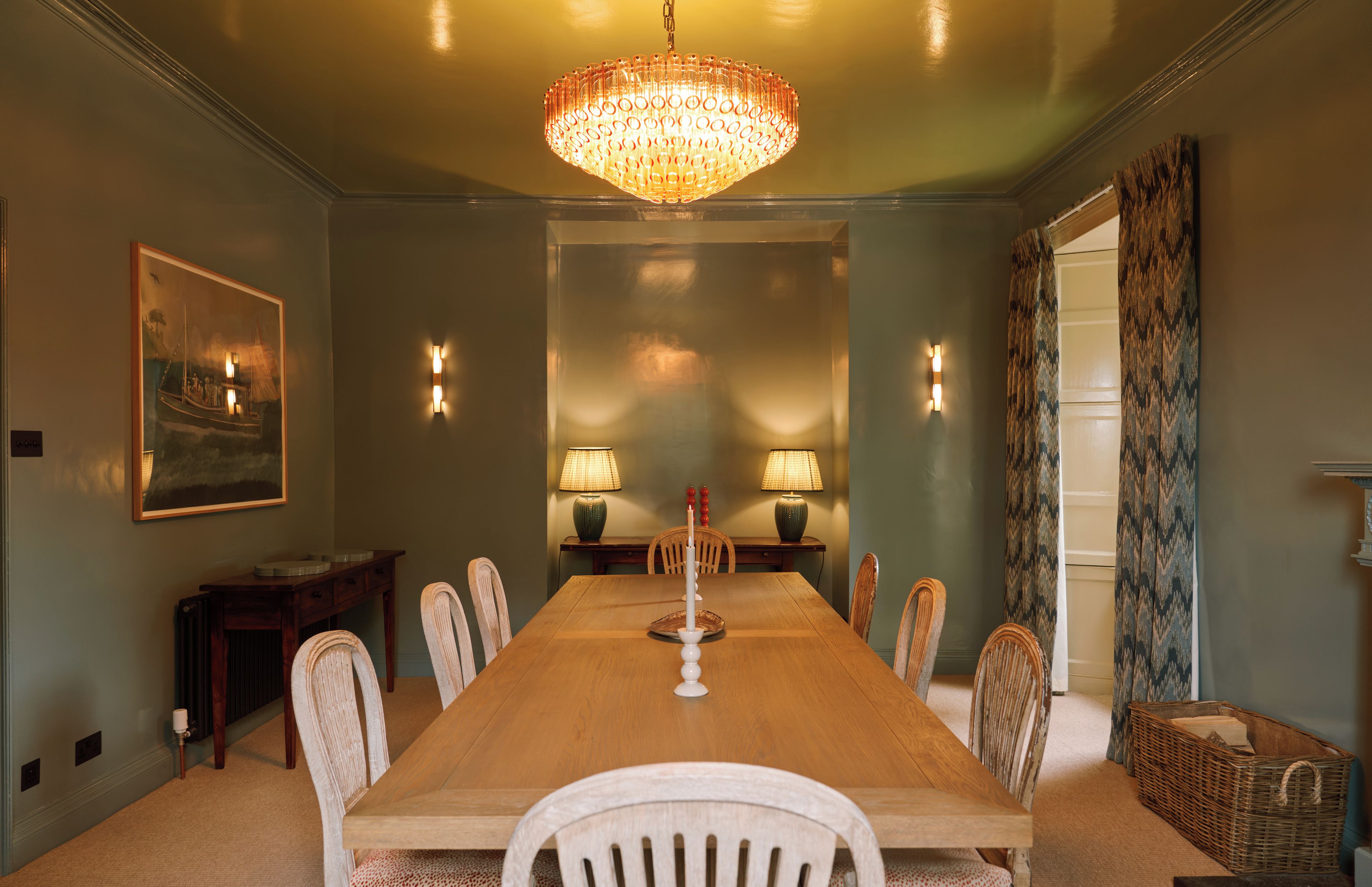 A dining room with a long wooden table, six chairs, two wall-mounted lamps, and a large chandelier.