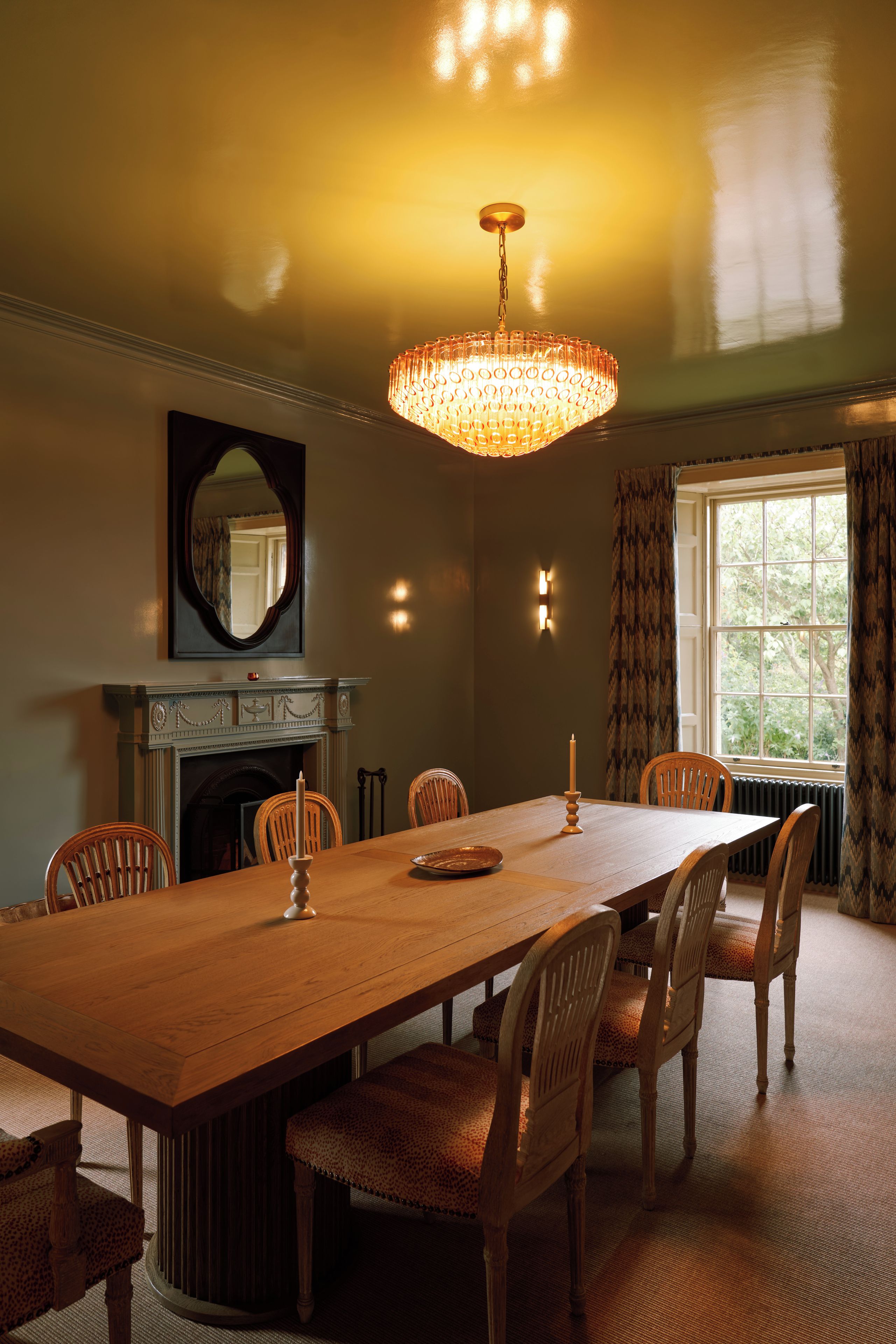 Een eetzaal met een lange houten tafel, zes stoelen, een kroonluchter, een open haard en een grote spiegel aan de muur.