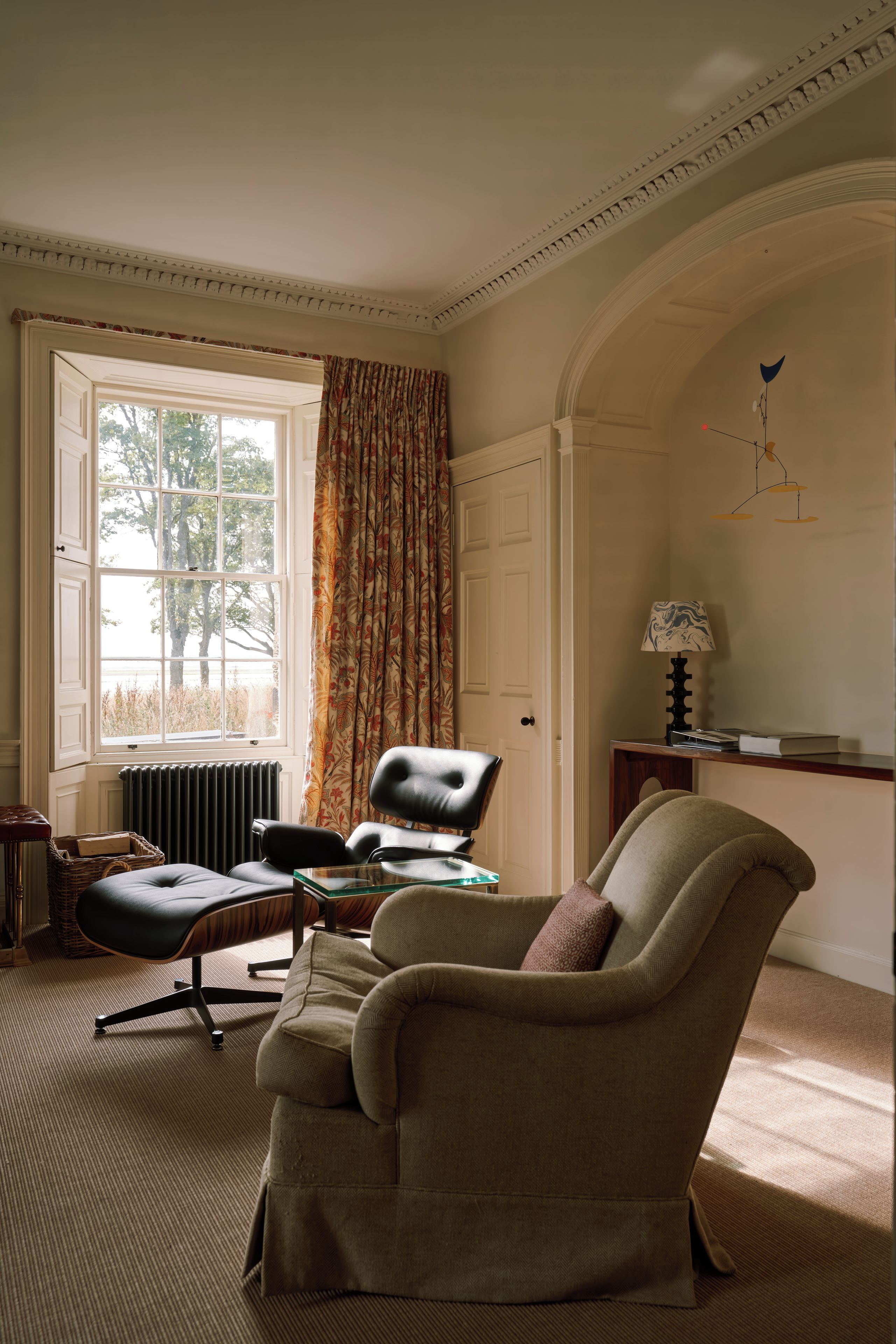 Een kamer met een groot raam, een radiator, een bureau met een lamp en twee stoelen.