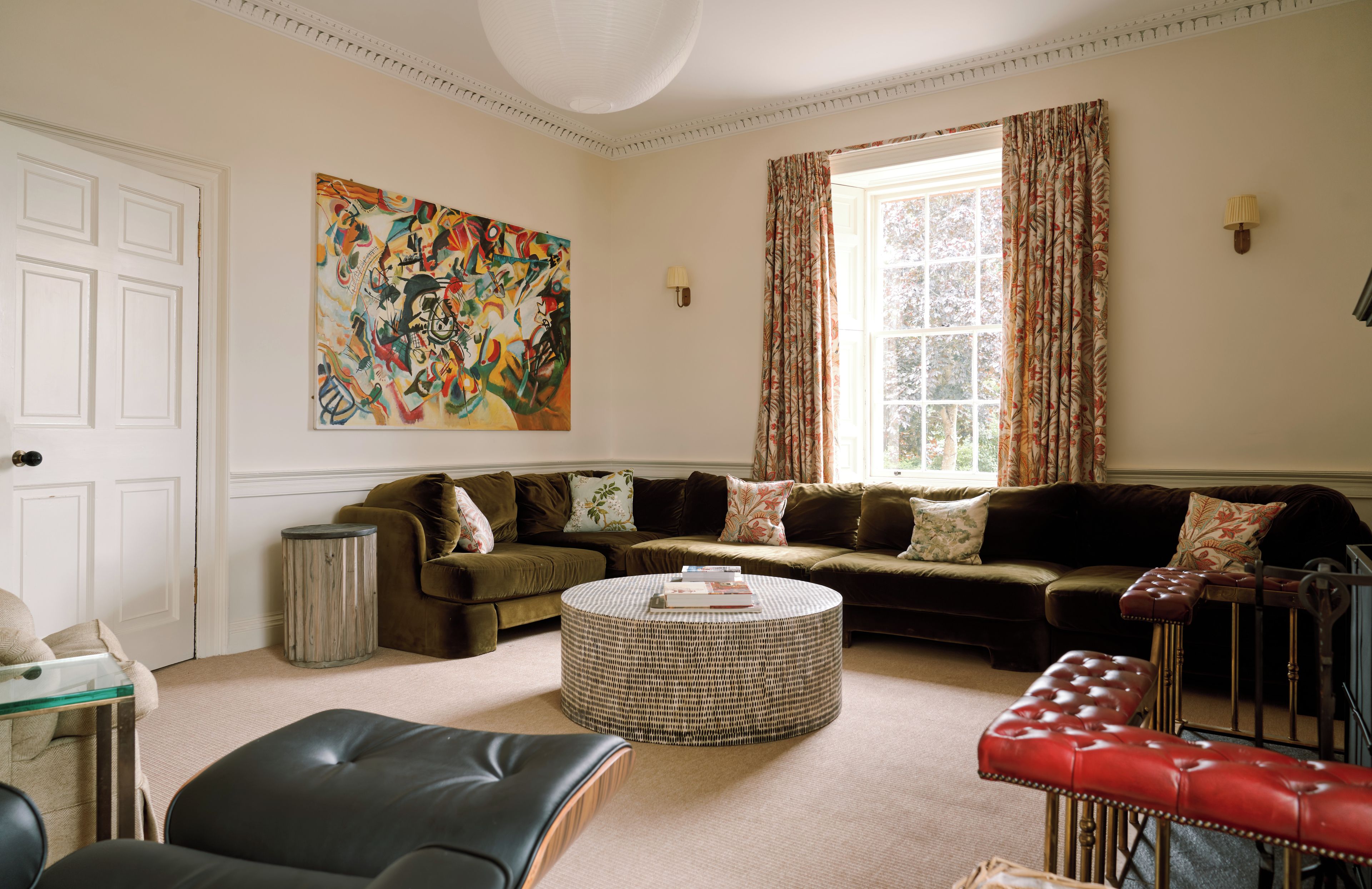 A living room with a sectional sofa, a round coffee table, a red leather ottoman, a large abstract painting, and a window with curtains.