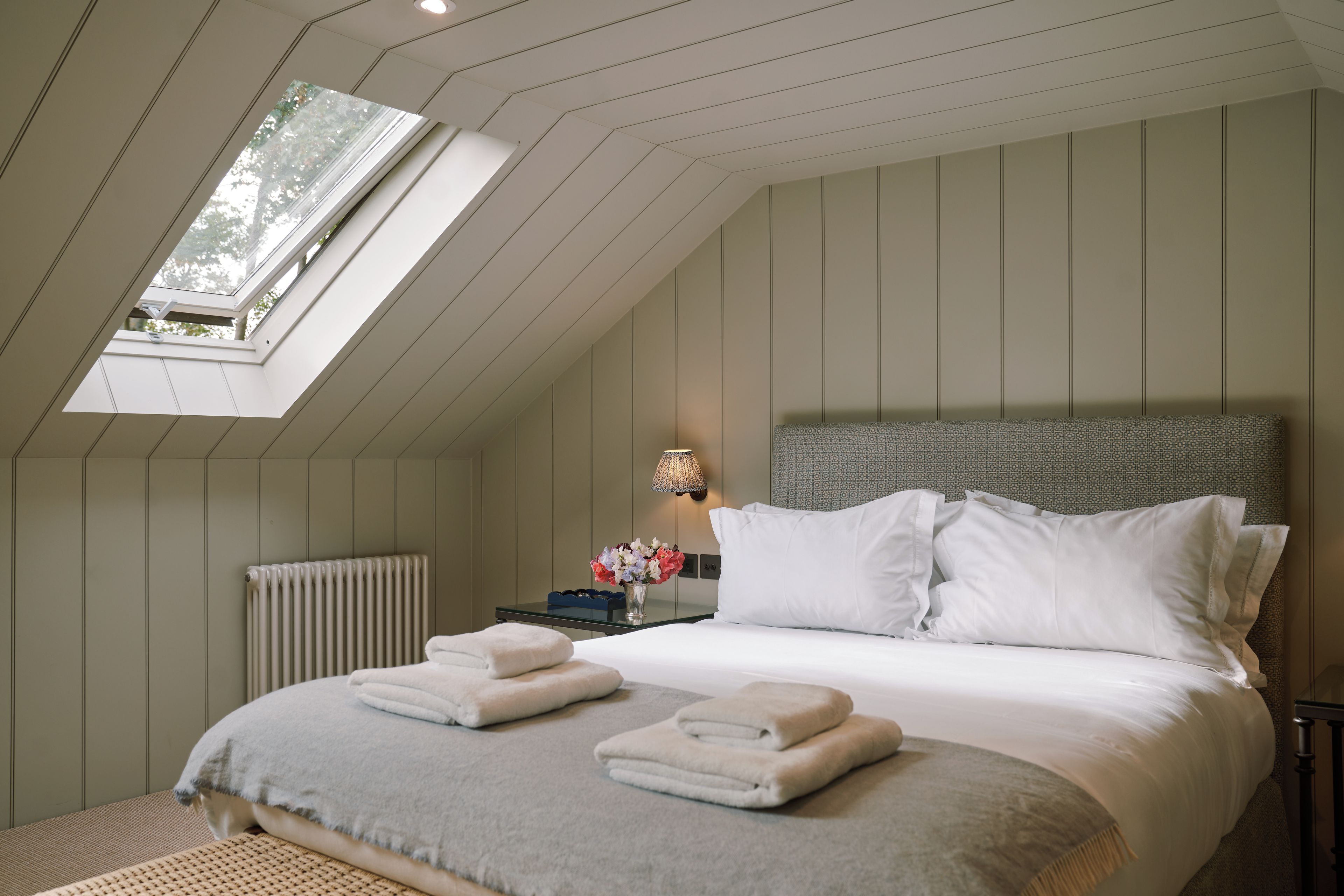A bedroom with a skylight, a bed with pillows, a bedside table with a lamp, and a radiator.