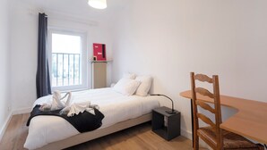 A bedroom with a bed, a bedside table, a chair, a window with curtains, and a radiator. - Hôtel des Cousines – Emma Apartment (Nancy)