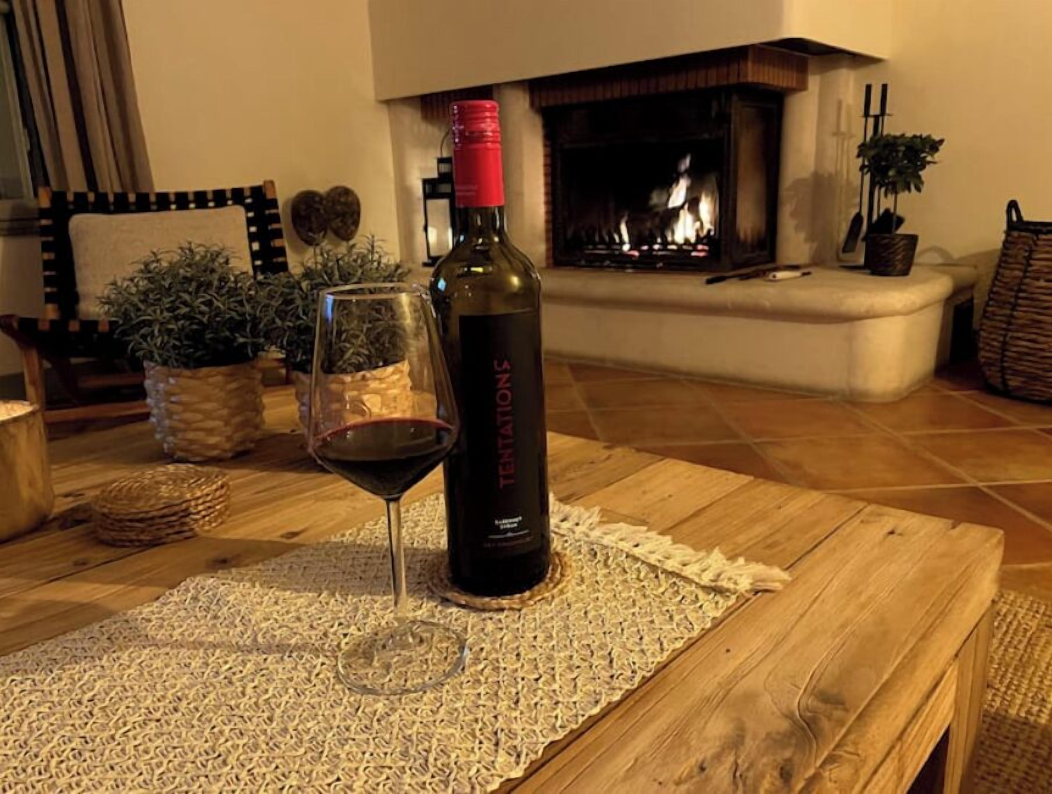 A bottle of wine and a glass on a wooden table in a cozy living room with a fireplace.