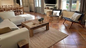 Amplia sala de estar con un sofá blanco, una mesa de centro de madera y un televisor montado en la pared. Hay dos sillones con cojines, un ventanal con cortinas y un suelo de baldosas.