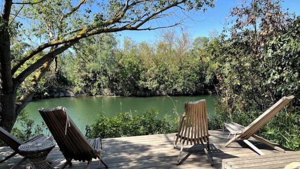 Una terraza de madera con dos sillones de descanso frente a una masa de agua tranquila, rodeada de exuberante vegetación.