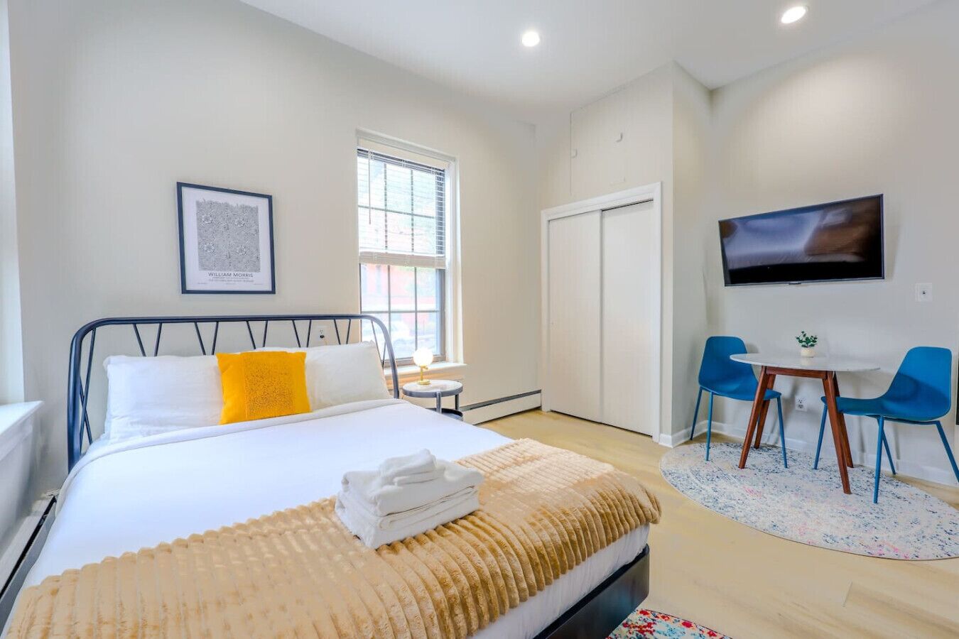 A hotel room with a bed, a desk with two chairs, a TV, and a window with blinds.