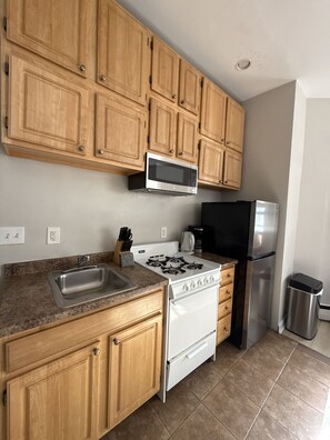 A kitchen with wooden cabinets, a stainless steel sink, a gas stove, a microwave, a refrigerator, and a trash bin. - Comfy Back Bay Alcove Studio Apt (Boston)