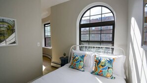 A bedroom with a large arched window, a bed with white bedding and colorful pillows, a small bedside table with a plant, and a framed picture on the wall. - Comfy Back Bay Alcove Studio Apt (Boston)