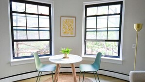 A room with a round table, two chairs, a rug, and a bed. There are two windows with blinds and a floor lamp.
